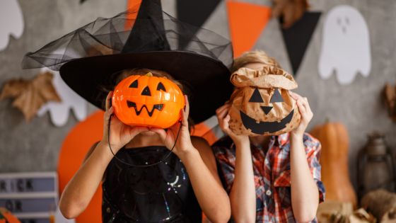 A halloween decorated background. In the centre are two children, one dressed in a black witch outfir, the other a checked shirt, perhaps a scarecrow. They are holding jack-o-lantern faces in front of their own faces.