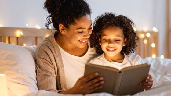 A photo of a black/mixed race mother sat in bed with her child. They are both smiling whilst looking at a book together. There are fairy lights in the background.