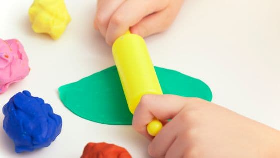 A side photo of a child's hands with a yellow rolling pin, rolling out some green playdough. There are also balls of yellow, pink, blue, and red playdough on the table.