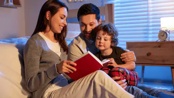Image of a family with a man, woman, and child, sitting together in a bedroom on the floor looking at a book.