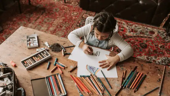 A child is sat drawing a picture at a wooden table that has lots of crayons and coloured pencils.
