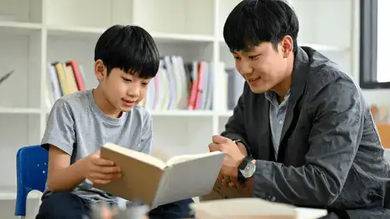 a father and son are sat together, maybe in a library, reading a book.