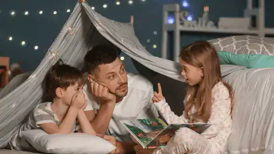 A father is sat in a den with his two children. The eldest, a girl, has a book in her hand and is talking to her family.