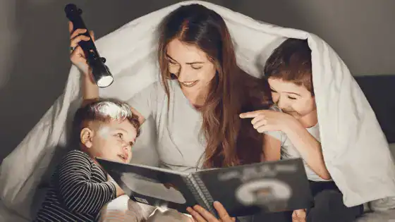 Mother and two children sat in a den with a torchlight reading a bedtime story together.