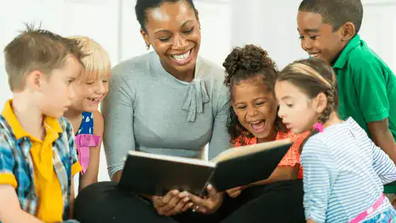 A teacher is sat holding a book and smiling whilst reading to students stood beside her