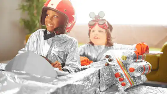 two boys sat in a pretend racing car with helmet and goggles on.