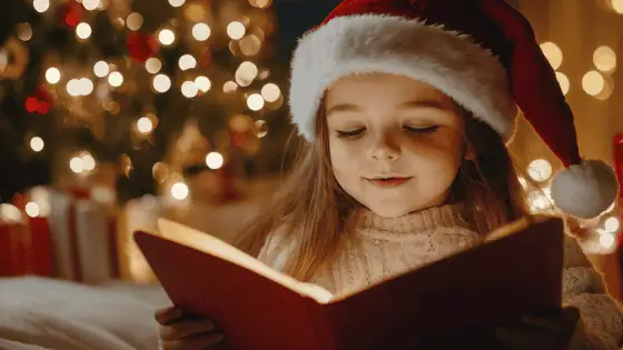 a young child with a santa hat on reading a story book in front of a christmas tree